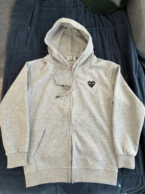 Grå hoodie från Comme des Garçons Play - Snygg grå hoodie från Comme des Garçons Play med dragkedja, huva och snörning. På bröstet sitter det ikoniska svarta hjärta-märket med ögon. Tillverkad i mjuk bomullsblandning och har två fickor framtill. Perfekt för en avslappnad och trendig look.