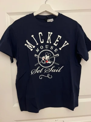 Marinblå Mickey Mouse t-shirt - Snygg marinblå t-shirt från H&M med stort Mickey Mouse-tryck och texten 'Set Sail' framtill. Klassisk passform och rund hals. Perfekt för dig som gillar retro och tecknade motiv.