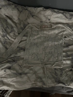 Grå sweatshirt med lama-mönster - Supersöt grå sweatshirt från Censored med tryck av tecknade lamor över hela tröjan. Tröjan har långa ärmar, rund halsringning och en knytning nedtill för extra detalj. Perfekt för dig som gillar gulliga djurmotiv och avslappnad stil.