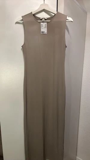 Beige långklänning från H&M - Stilren beige långklänning från H&M i ett mjukt och stretchigt material. Klänningen är ärmlös med rund halsringning och har en rak, enkel siluett som ger en modern look. Perfekt för dig som gillar minimalistisk stil.