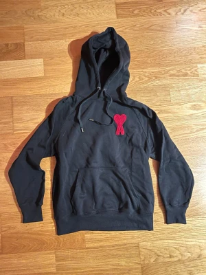 Svart hoodie med rött hjärtmärke - Säljer en svart hoodie från AMI Paris med deras ikoniska röda hjärtmärke broderat på bröstet. Hoodien har dragsko i huvan och en stor magficka. Tillverkad i mjuk bomullsblandning och har ribbade muddar vid ärmar och nederkant. Perfekt för en avslappnad streetwear-look.
