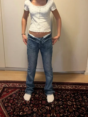 Blå low waist jeans med v-formadnning och ett linne till  - Säljer ett par blå low waist jeans med unik v-formad linning framtill och snygga detaljerade bakfickor. Jeansen har raka ben och klassisk femficksdesign. Perfekta för dig som gillar en trendig och avslappnad stil. Linné med små knappar och ett svart vitt prickigt mönster