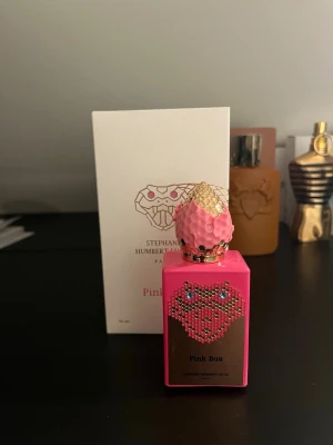 Pink Boa parfym från Stephane Humbert Lucas - Unik parfymflaska i starkt rosa med gulddetaljer och gnistrande stenar. Flaskan har en dekorativ, texturerad kork och en elegant orm-design på framsidan. Lyxig och iögonfallande för dig som gillar exklusiva dofter.