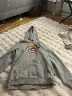 Grå Dickies hoodie med logga - Säljer en grå hoodie från Dickies med stor färgglad logga på bröstet. Tröjan har huva och en stor magficka framtill. Klassisk passform och mjukt material som är perfekt för chill dagar. Ribbade muddar vid ärmslut och nederkant. Storlek L men passar M!!!