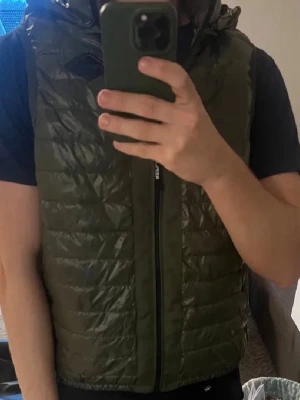 Olivgrön dunväst från Replay - Snygg olivgrön dunväst från Replay med quiltad design och hög krage. Västen har dragkedja framtill och två sidofickor. Perfekt att slänga över en hoodie eller t-shirt för extra värme, perfekt för våren!