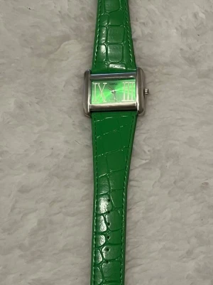 Grön rektangulär klocka med läderband - Stilren klocka från Swiss med rektangulärt silverfärgat boett och grön urtavla. Klockan har stora romerska siffror och ett grönt band med krokodilpräglad yta. Perfekt för dig som vill sticka ut med en färgglad accessoar. Tyvärr behöver klockan nytt batteri.