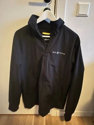 Svart vindjacka från Sail Racing - Snygg svart vindjacka från Sail Racing med hög krage och dragkedja framtill. Jackan har logga på bröstet och en grafisk detalj på ryggen. Tillverkad i polyester och har lång ärm. Perfekt för dig som gillar stilren och sportig look.
