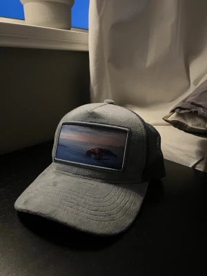 Ljusblå keps från Revenite - Säljer en oanvänd keps från Revenite med ett broderat motiv framtill som föreställer en sportbil i ett  landskap med solnedgång. Kepsen har böjd skärm och justerbar snapback. 