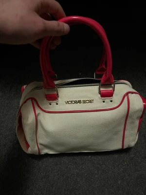 Victoria's Secret Vintage Väska - Snygg handväska från Victoria's Secret i vit canvas med rosa detaljer och handtag i glansigt material. Väskan har guldfärgad logga framtill och dragkedja upptill. Perfekt för dig som vill ha en trendig och rymlig väska med coola kontraster.