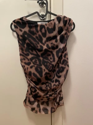Leopardmönstrad topp från Next - Säljer en leopardmönstrad topp från Next i tunnt, lätt material. Toppen har ärmlös design och en draperad, figurnära passform som ger en cool och trendig look. Perfekt för dig som vill sticka ut med ett djurmönster.