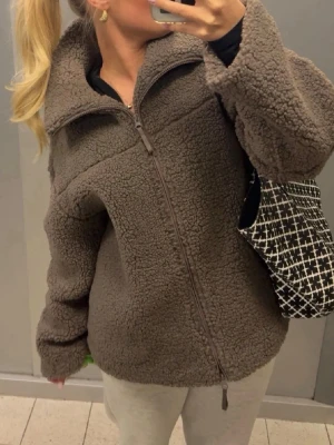 !lånad bild! Brun teddyjacka med dragkedja - Mysig brun teddyjacka med hög krage och hel dragkedja framtill. Jackan har en oversized passform och är gjord i fluffigt, mjukt material som ger en trendig look. Perfekt för att hålla värmen under kyliga dagar.