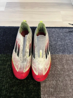 Adidas vita och röda fotbollsskor - Säljer ett par Adidas fotbollsskor utan snörning i vitt med röda och gula detaljer samt svarta ränder på sidorna. Skorna har en strömlinjeformad design och insida i limegrönt. Perfekta för dig som vill ha stilrena och snabba skor på planen.