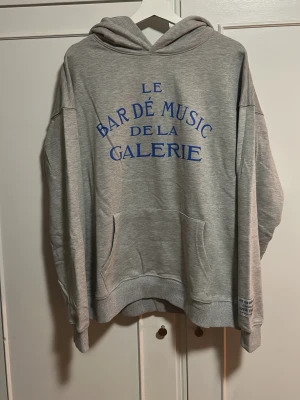 Grå hoodie Le Bar De Music Galerie (Gallery Dept) - Snygg grå hoodie från Gallery Dept med blå text 'Le Bar De Music De La Galerie' på bröstet. Hoodien har en stor huva, magficka och ribbade muddar. Tillverkad i mjuk bomullsblandning för extra komfort. Perfekt för en avslappnad och trendig look.