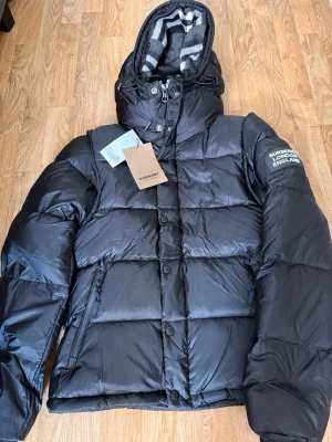  Burberry jacka  - Snygg svart pufferjacka från Burberry med huva och klassiskt rutigt foder. Jackan har dragkedja och knappar framtill, två sidofickor med dragkedja och Burberry-logga på ärmen. Perfekt för kalla vinterdagar och riktigt trendig look. Kan bli till en väst storlek S helt ny använd ett par gånger 