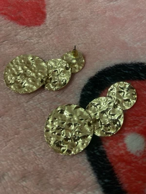 Guldiga örhängen med hamrad look - Säljer ett par statement örhängen i guldfärgad metall med tre runda, hamrade plattor i olika storlekar. Örhängena har stift och plastbaksida. Perfekta för dig som gillar unika accessoarer med lite extra edge en plopp saknas från ett örhänge .