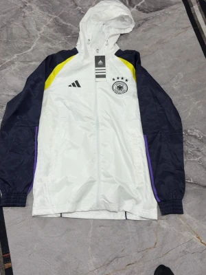 Adidas vindjacka Tyskland vit/svart - Snygg vindjacka från Adidas med Tysklands landslagslogga. Jackan är vit med svarta ärmar, gula detaljer och lila inslag. Den har huva, dragkedja och elastiska muddar. Perfekt för sportiga dagar och coola outfits.