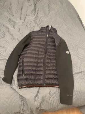 Svart pufferjacka från Moncler - Snygg svart pufferjacka från Moncler med quiltad kropp och släta svarta ärmar. Jackan har hög krage, dragkedja framtill och en ficka med dragkedja på sidan. Moncler-logga på ena ärmen och diskret logotyp vid nederkanten.