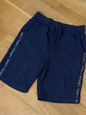 Marinblå shorts Tommy Hilfiger - Snygga marinblå shorts från Tommy Hilfiger med logoband längs sidorna och dragsko i midjan. Mjukt material och två fickor framtill. Perfekta för snygga sommardagar eller chill hemma. 