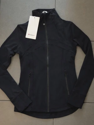 Svart vindjacka från Lululemon - Slimmad svart vindjacka från Lululemon med hög krage och hel dragkedja framtill. Jackan har långa ärmar och två fickor med dragkedja. Perfekt för dig som vill ha en sportig och stilren look.