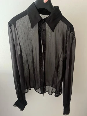 Svart transparent skjorta med knappar - Snygg svart transparent skjorta med tunna vertikala ränder och klassisk krage. Skjortan har långa ärmar och knappar framtill. Perfekt för dig som vill ha en edgy och trendig look.