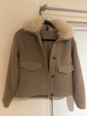 Beige kappa med fuskpäls - Stilren beige kappa med stor krage i ljus fuskpäls och två stora fickor framtill. Kappan har raka ärmar, knäppning med stora knappar och är tillverkad i ett mjukt ull-liknande material. Perfekt för dig som vill ha en trendig och mysig look.