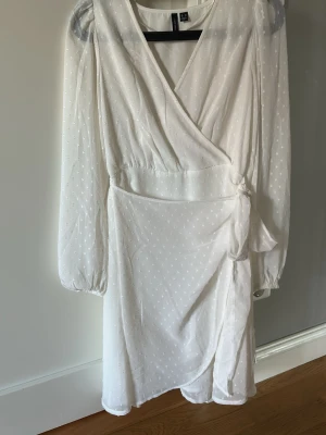 Vit omlottklänning med knyt - Säljer en vit kort omlottklänning med knyt i midjan och diskret prickigt mönster. Klänningen har långärmade, lätt transparenta ärmar och v-ringad hals. Perfekt för sommarens alla tillfällen!