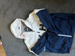 Helly Hansen blåvit vindjacka - Snygg blåvit vindjacka från Helly Hansen med stor logga på ryggen och mindre HH-logga på huvan. Jackan har dragkedja framtill, justerbar huva och elastiska muddar. Perfekt för blåsiga dagar och streetwear-stil.