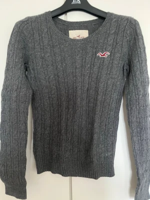 Grå kabelstickad tröja Hollister - Snygg grå kabelstickad tröja från Hollister med rund halsringning och diskret logga i rött och vitt på bröstet. Tröjan har långa ärmar och ribbade muddar vid ärmslut och nederkant. Perfekt för lager på lager och enkel att matcha.