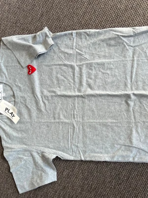 Grå Comme des Garçons PLAY t-shirt - Säljer en grå t-shirt från Comme des Garçons PLAY med det ikoniska röda hjärtat broderat på bröstet. T-shirten har korta ärmar, rund hals och är gjord i mjuk bomull. Perfekt basic med en twist för dig som gillar streetwear vibes.