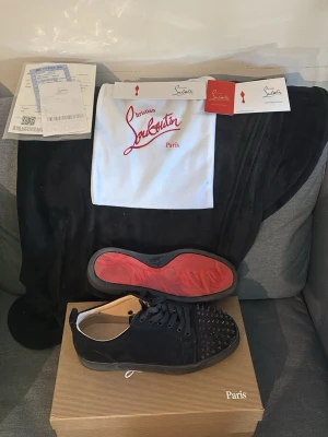 Svarta Christian Louboutin sneakers - Säljer ett par svarta sneakers från Christian Louboutin  i storlek 41 med klassisk röd sula och coola nitar på tån. Skorna har snörning samt ovandel i mocka. Perfekta för dig som vill sticka ut med exklusiv design och edgy detaljer. Skorna är knappt använda. Det ingår en Louboutin kartong, skopåse och Louboutin kort.
