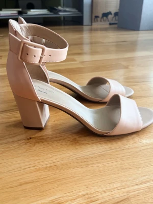 Babyrosa klackar från Clarks - Säljer ett par stilrena pumps från Clarks i nude/rosa färg med bred klack och öppen tå. Skorna har en bred rem över tårna och justerbart ankelband med spänne. Tillverkade i slätt läderliknande material, perfekta till många olika outfits. Väldigt fina hade gärna behållt dem själv om det inte vore att de var en aning för stora, står strl 38 men tycker de är mer 38,5, i princip nya använt en gång. 