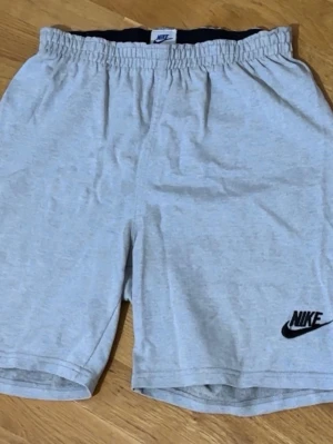 Grå Nike shorts med logga - Snygga ljusgrå shorts från Nike med svart logga på vänster ben. De har elastisk midja för skön passform och är tillverkade i mjuk bomull, perfekta för chill dagar eller träning.