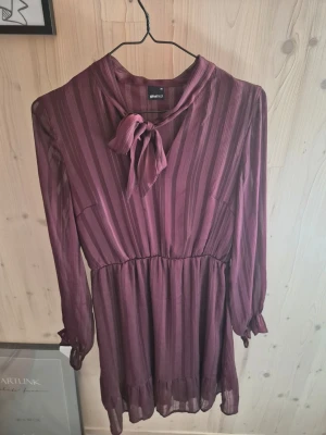 Vinröd randig klänning från Gina Tricot - Vinröd kortklänning från Gina Tricot med vertikala ränder, knytband i halsen och långa ärmar med lätt volang vid ärmslut. Tillverkad i ett tunt, flowy material som ger en elegant look. Perfekt för dig som gillar feminina detaljer och stilren design.