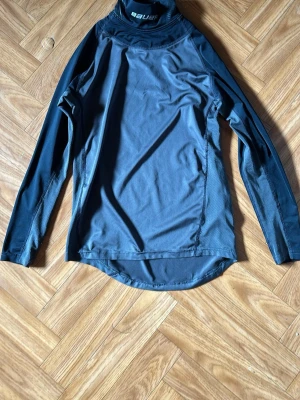 Svart funktions-tröja från Bauer - Säljer en svart långärmad funktions-tröja från Bauer med hög krage och logga på halsen. Tillverkad i ett stretchigt och snabbtorkande material, perfekt för träning eller sportaktiviteter. Tröjan har en sportig passform och ventilerande paneler. Men inbyggd halsskydd för hockeyn. Med storlek M/M youth.