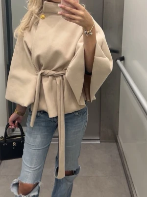 Poncho kappa  - Stilren beige poncho kappa. Kappan har hög krage med guldfärgad knappdetalj och är tillverkad i ett mjukt tyg som ger en lyxig känsla. Perfekt för dig som vill ha en trendig och elegant look. Från Missäy 