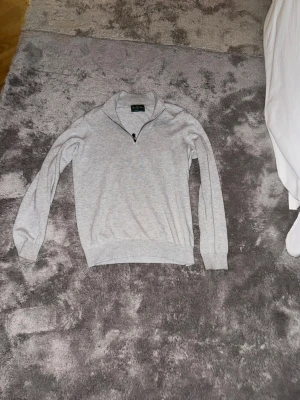 Ljusgrå stickad tröja från Alan Paine - Ljusgrå stickad tröja från Alan Paine i mjuk bomull. Tröjan har halv dragkedja vid halsen, ribbade muddar och en klassisk passform. Perfekt för lager på lager och enkel att matcha med olika outfits. Köpt från nk.