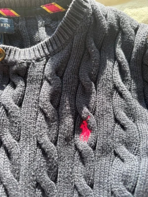 Mörkblå kabelstickad kofta Polo Ralph Lauren - Kabelstickad mörkblå kofta från Polo Ralph Lauren med rund halsringning och knappar framtill. Klassisk röd logga broderad på bröstet. Långa ärmar och ribbade muddar. Perfekt för lager på lager och en tidlös stil.