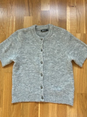 Grå kortärmad kofta Gina Tricot - Mysig gråmelerad kofta från Gina Tricot med korta ärmar och rund hals. Koftan har knappar framtill och ribbade muddar vid ärmslut och nederkant. Perfekt att slänga över en topp för en chill look.