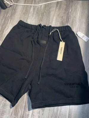 Svarta shorts från Essentials Fear of God - Snygga svarta shorts från Essentials Fear of God med långa snören i midjan och diskret logga på benet. Tillverkade i mjuk bomull för en bekväm känsla. Perfekta för en avslappnad och trendig stil. skicka 10/10 aldrig använda