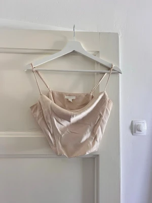 Champagnefärgad satin linnetopp - Snygg croppad linnetopp i champagnefärgad satin med tunna axelband och draperad urringning i strl M (passar en S). Perfekt för dig som gillar minimalistisk och trendig stil. Enkel att matcha med både jeans och kjol.