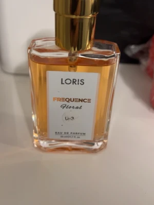 Loris Frequence Floral parfym - Loris Frequence Floral Eau de Parfum i en elegant, rektangulär glasflaska med guldlock. Doften är blommig och flaskan rymmer 50 ml. Etiketten är vit med guld- och svart text som ger en lyxig känsla.