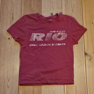Vinröd t-shirt med Rio de Janeiro-tryck - Magenta färgad t-shirt med stort vitt tryck på bröstet där det står 'Brazil Rio de Janeiro' i en sprucken stil, den var köpt så, inget som hänt av slitning. 