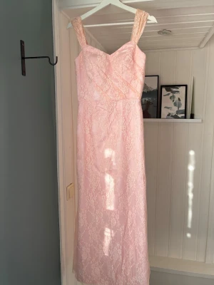 Ljusrosa spetsklänning JJ's House - Söt ljusrosa långklänning från JJ's House med breda axelband och vacker spetsdetalj över hela klänningen. Klänningen har en figurnära passform upptill och är fodrad med mjukt tyg. Perfekt för bal, student eller festliga tillfällen.