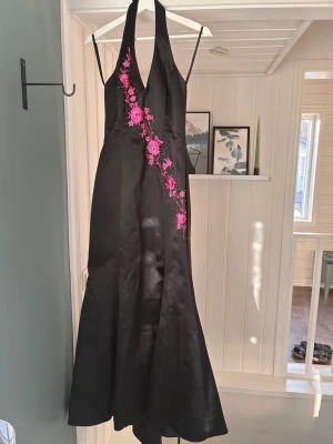 Svart klänning med rosa broderi Gino Cerruti - Säljer en elegant svart klänning från Gino Cerruti med halterneck och v-ringad front. Klänningen har rosa broderade blommor längs ena sidan och är figurnära upptill med utsvängd nederdel. Perfekt för dig som vill sticka ut med snygga detaljer.