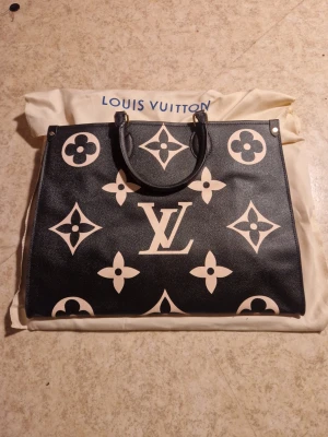 Louis Vuitton svart handväska - Snygg svart handväska från Louis Vuitton med stora beige monogram och klassiska LV-detaljer. Väskan har två handtag och röd textilinsida med dragkedjefack. Tillverkad i Frankrike och levereras med dustbag. Perfekt för dig som vill ha en statementväska.