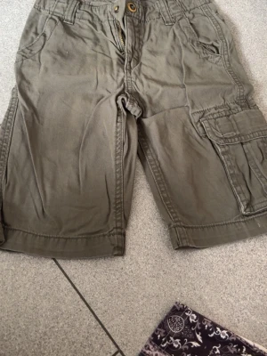 Gröna cargoshorts från H&M L.O.G.G. - Snygga gröna cargoshorts från H&M L.O.G.G. med flera fickor och klassisk cargomodell. Shorts med knapp och dragkedja framtill samt praktiska sidofickor och benficka. Perfekta för varma dagar och enkel att matcha med t-shirt.