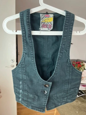 Clubs väst - Snygg jeansväst från Cheap Monday med två knappar framtill och dekorativa fickor. Västen har en justerbar knytning i ryggen och cool tvättad denimlook.                                     Storlek M men passar som S                                   Axelbredd 24 cm                                                       Längd 48 cm