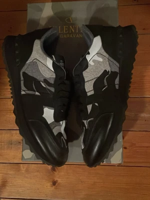 Valentino Garavani camo sneakers - Säljer ett par snygga sneakers från Valentino Garavani med svart, grå och vit camomönstrad ovandel. Skorna har svarta snören, robust sula och detaljer i läder och textil. Perfekta för dig som vill sticka ut med en cool streetwear-look.