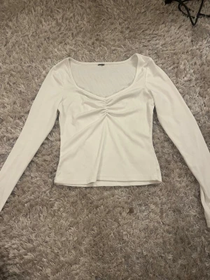 Vit ribbad långärmad topp - Snygg vit långärmad topp med ribbad struktur och v-ringning. Toppen har en lätt rynkning framtill som ger en fin detalj och sitter figurnära. Perfekt att styla med jeans eller kjol för en trendig look.