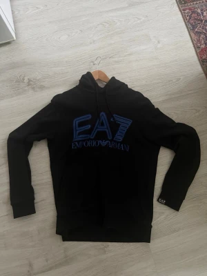 Svart EA7 hoodie från Emporio Armani - Svart hoodie från Emporio Armani EA7 med stor blå logga framtill. Klassisk huva med dragsko och ribbade muddar. Snygg och stilren design, perfekt för dig som gillar streetwear och exklusiva märken.